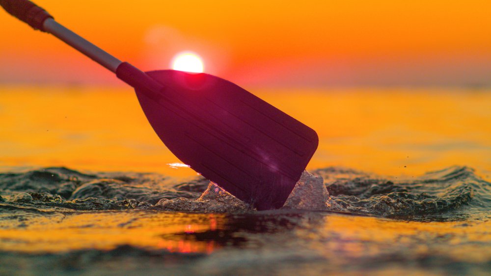 Oar in Sunset Waters Close-up of a kayak paddle slicing through water at sunset, with vibrant orange hues reflecting a peaceful and adventurous moment on the water.